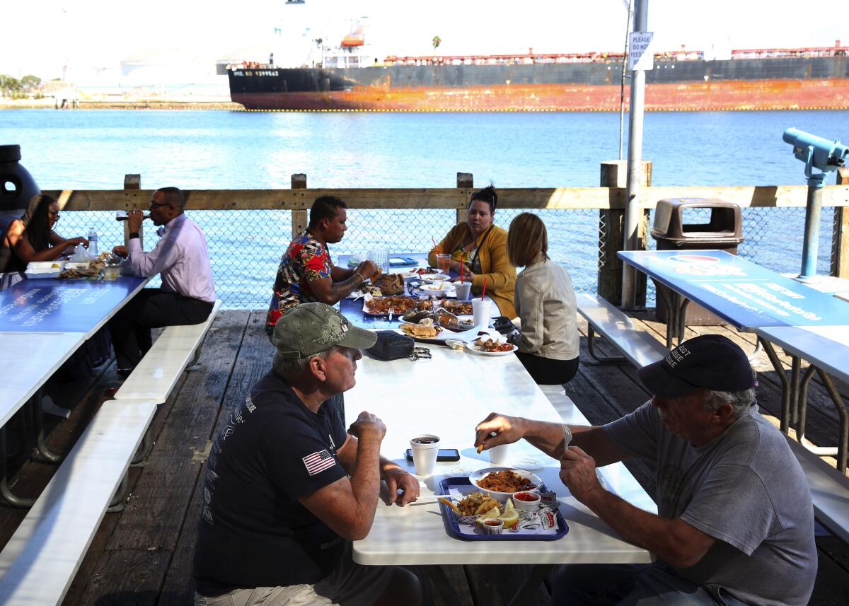 People eat outside, on the patio of the San Pedro Fish Market and Restaurant. A large ship is docked nearby across the bay.