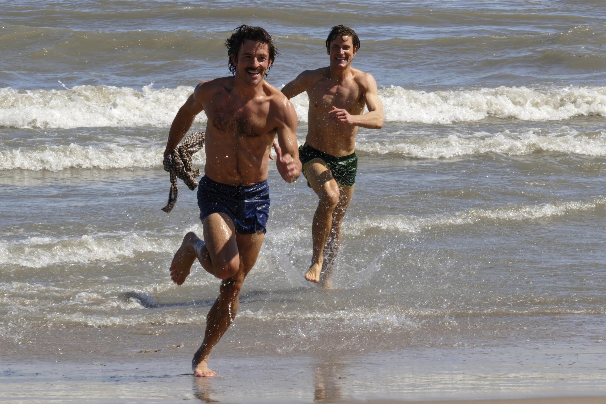 Two bare-chested men in swimming trunks run through the waves on the beach.