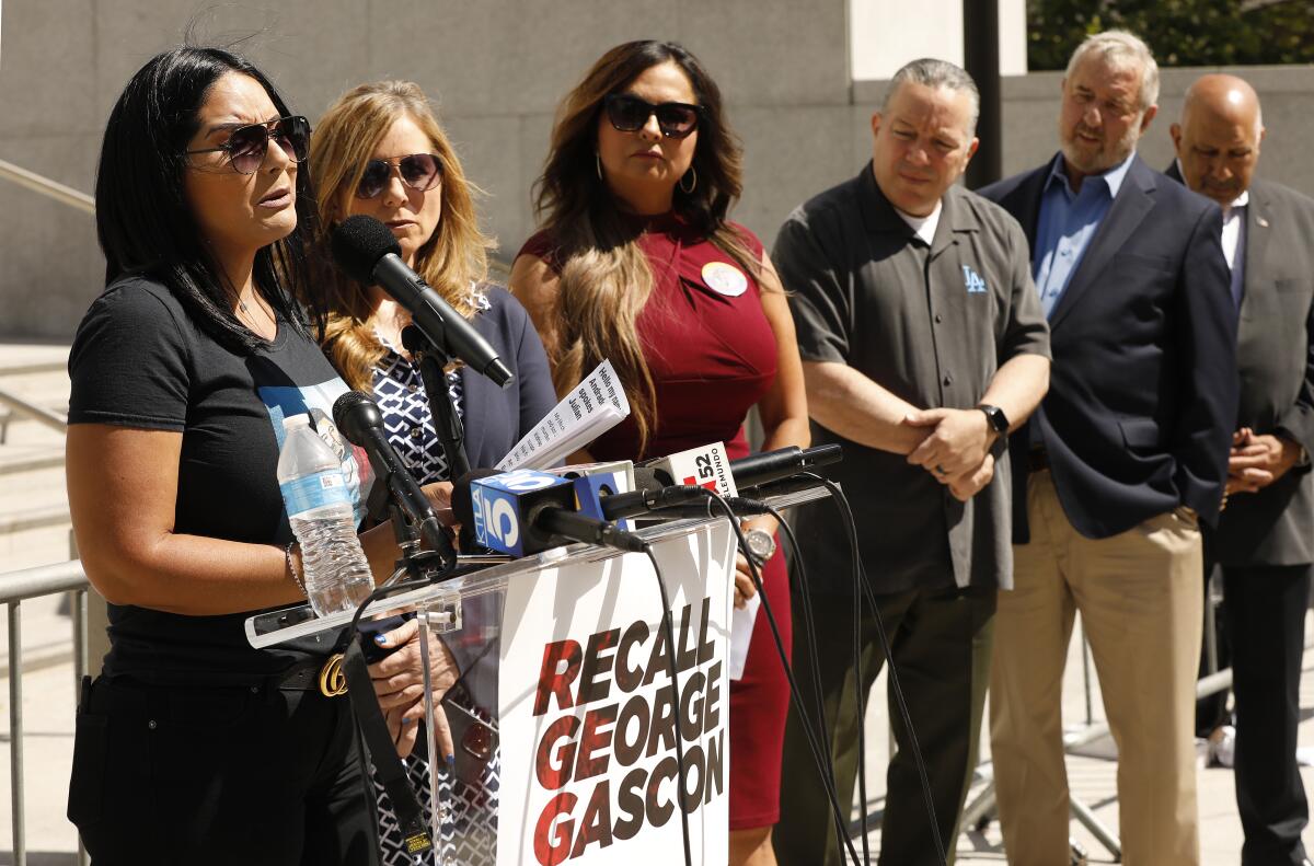 A woman standing with a small group of people talks at a lectern with a sign reading "Recall George Gascón."
