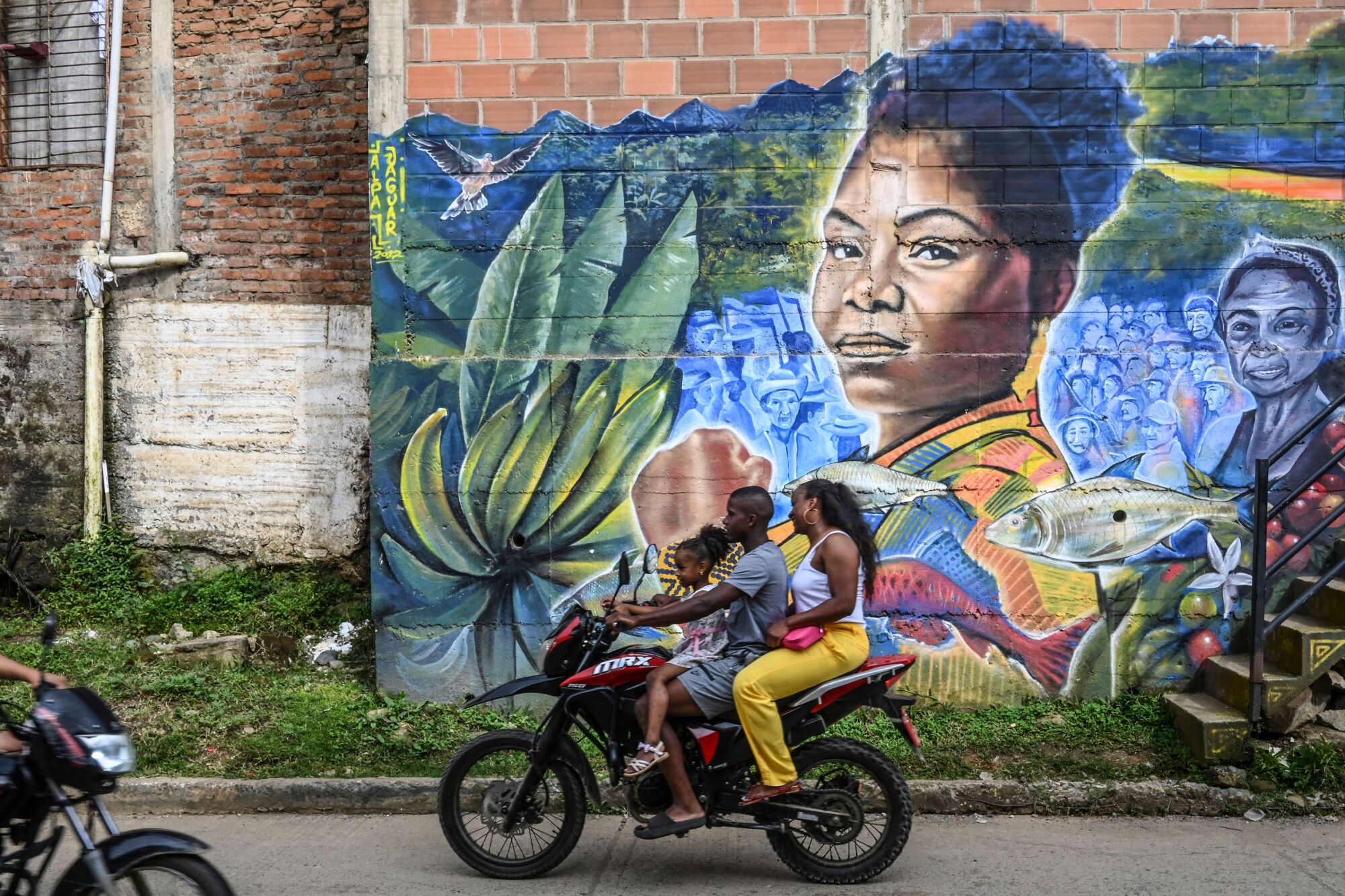 Three people on a motorcycle pass a mural of a woman's face