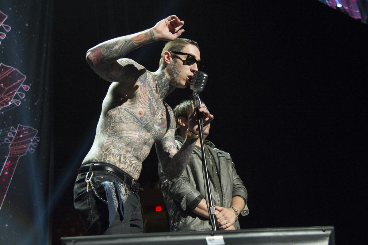 A shirtless Trace Cyrus, who is covered in body tattoos, speaks into a microphone onstage