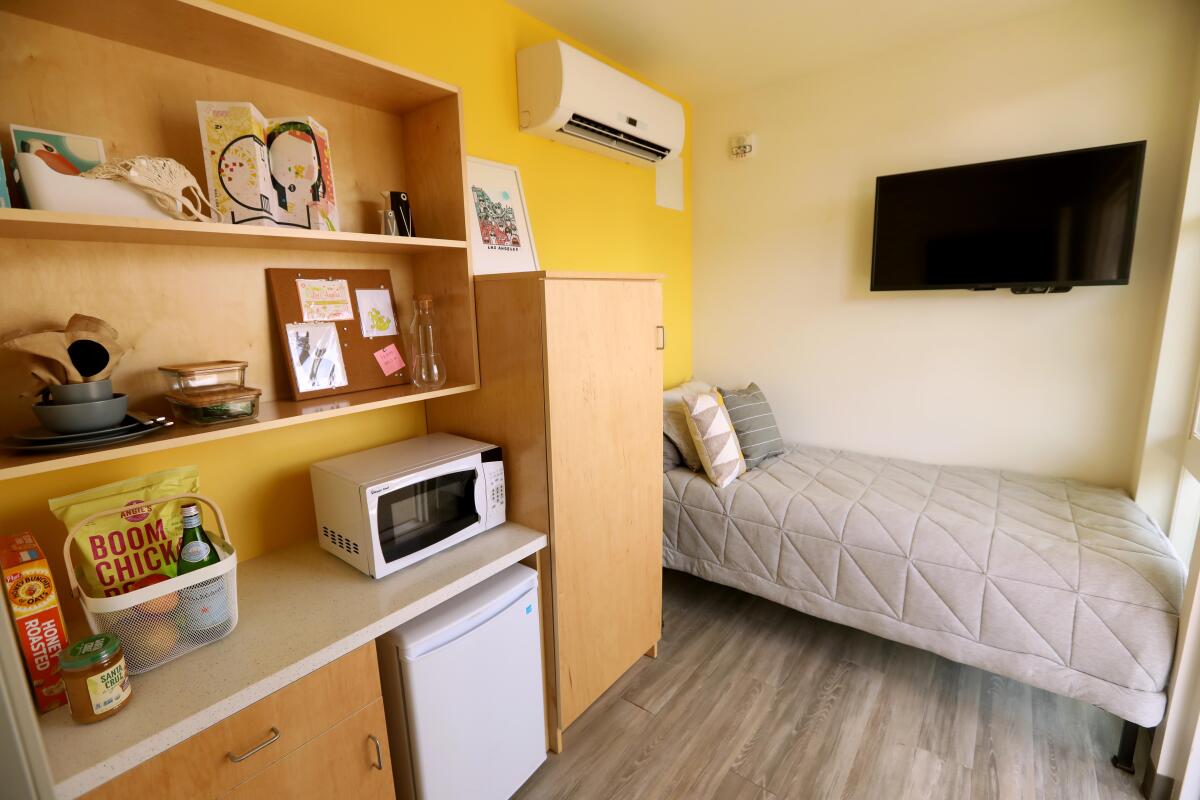An interior view of a shipping container apartment shows a tidy room with a mustard-colored wall and a single bed