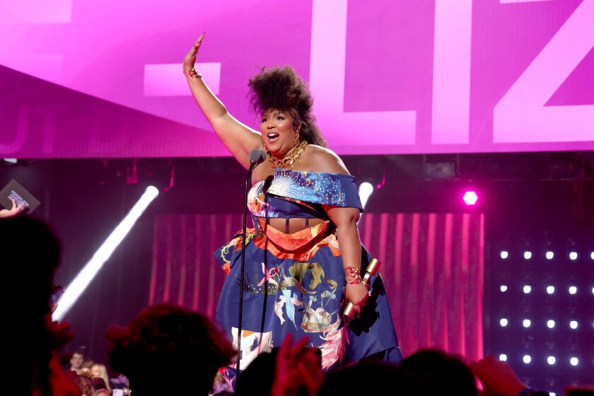 A woman in a colorful gown raises her right hand onstage while holding an award in the other