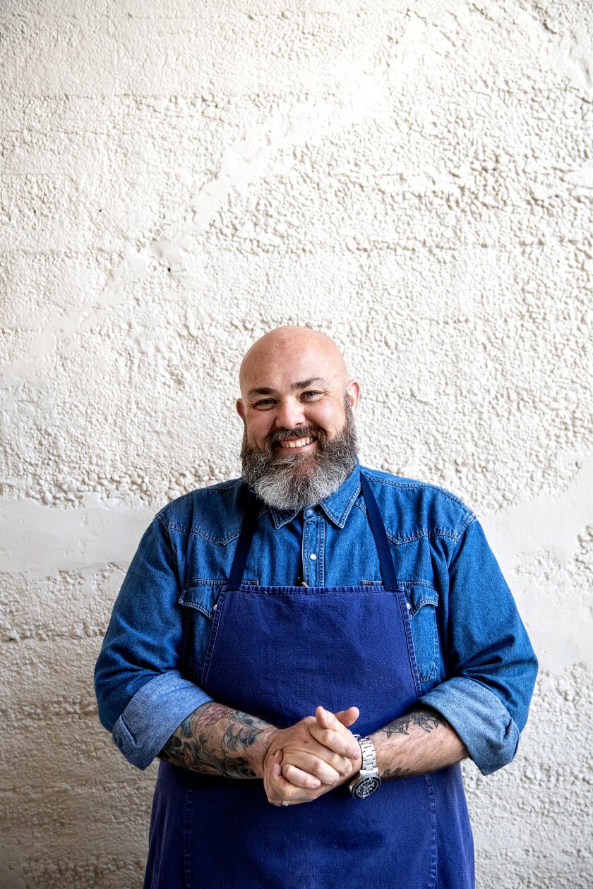 A bearded man in a blue apron and denim shirt stands against a white wall.