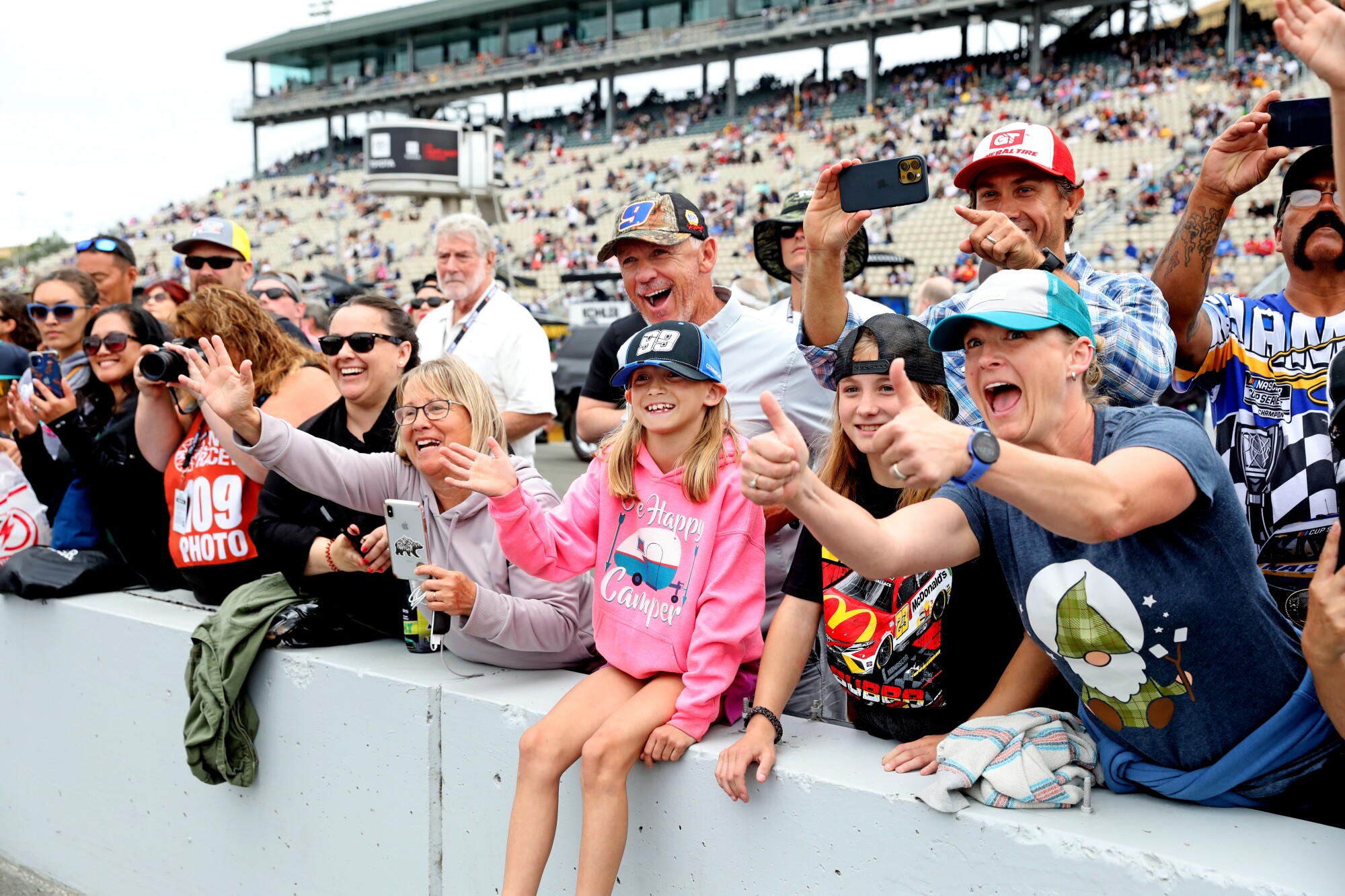 Jocee Holmes of Klamath Falls, Ore., wears a No. 99 car cap while sitting on a barrier and waving to Daniel Suarez.