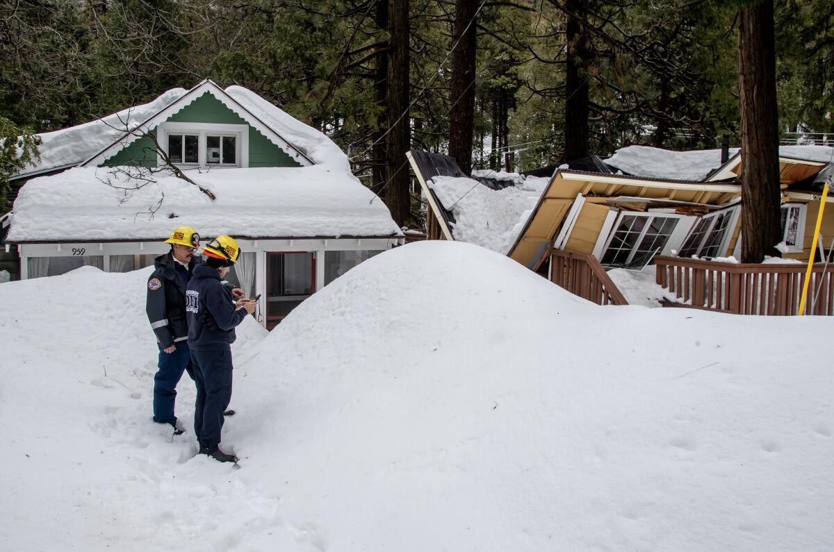 Two men in uniform with yellow helmets stand amid piles of snow and evergreens next to a damaged home.