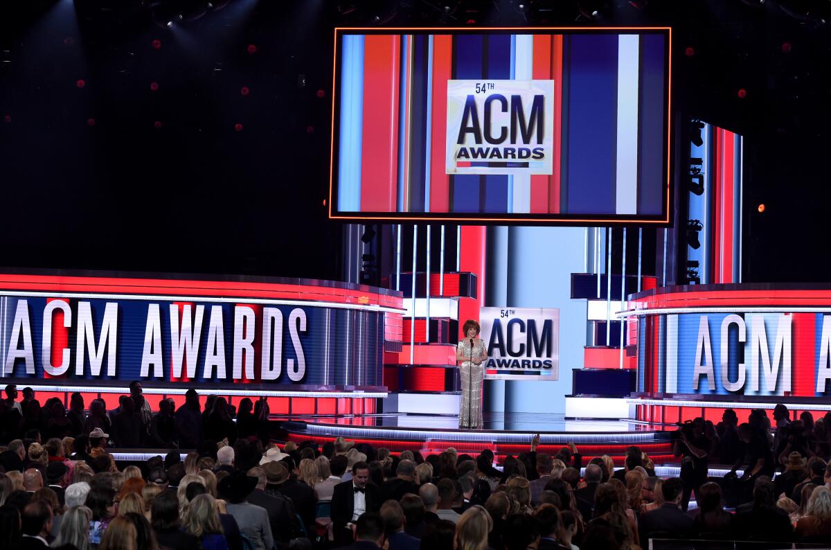 A woman in formal attire stands at a microphone in front of screens that say ACM Awards.