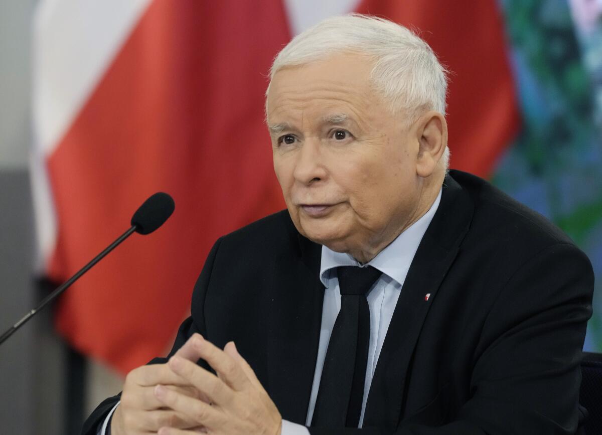  A man in a dark suit and tie is seated with his hands clasped before a microphone 