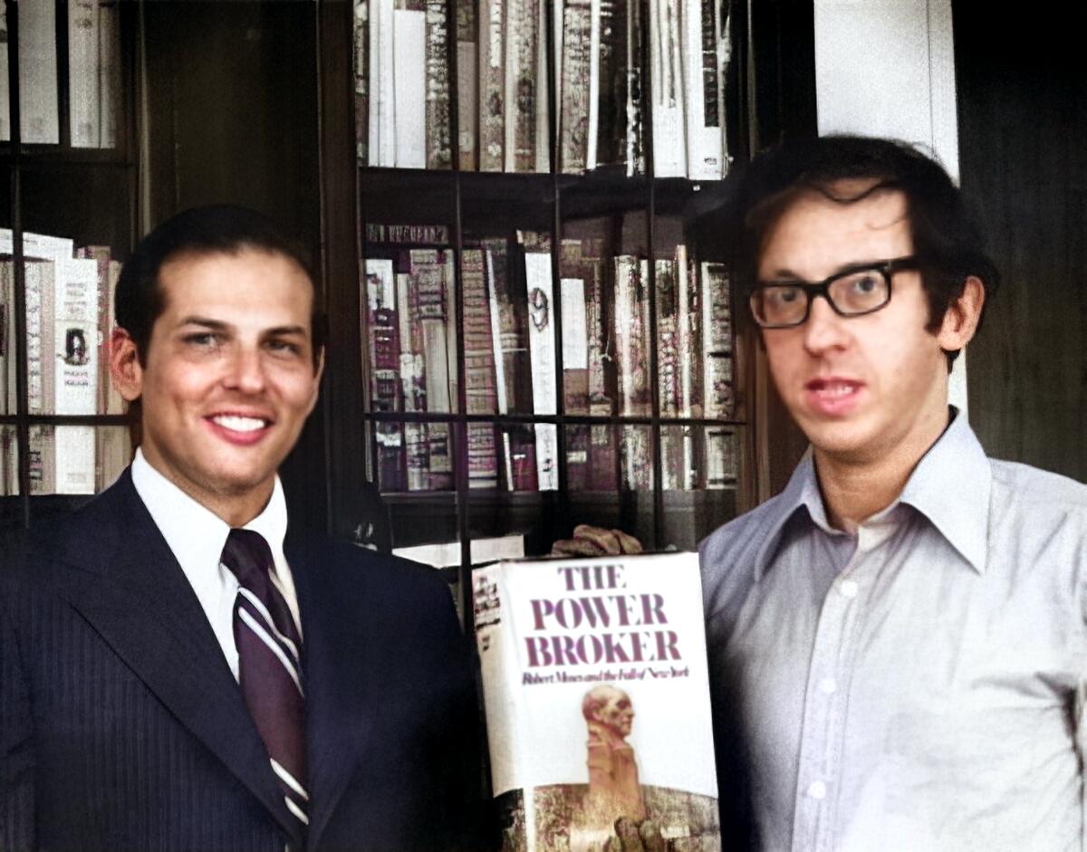 A smiling man in a suit and tie and a second in a dress shirt, with a copy of the book "The Power Broker." 
