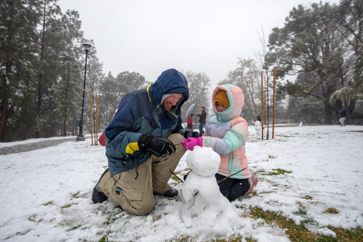 A man and child kneel down as they build a snowman in a park.