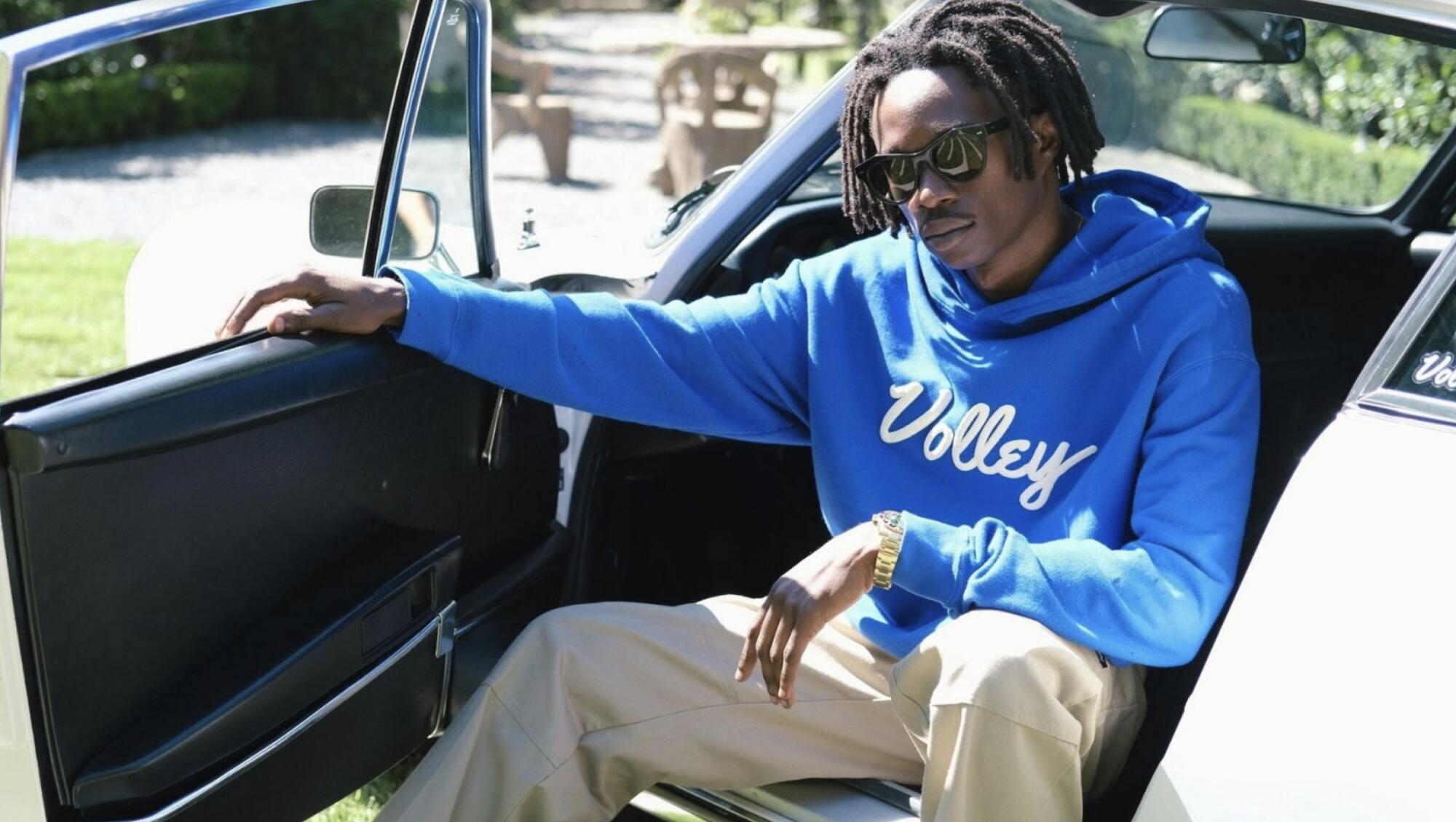 A man sits in a convertible car with the door open, wearing a blue hoodie with the word "Volley" in white script.