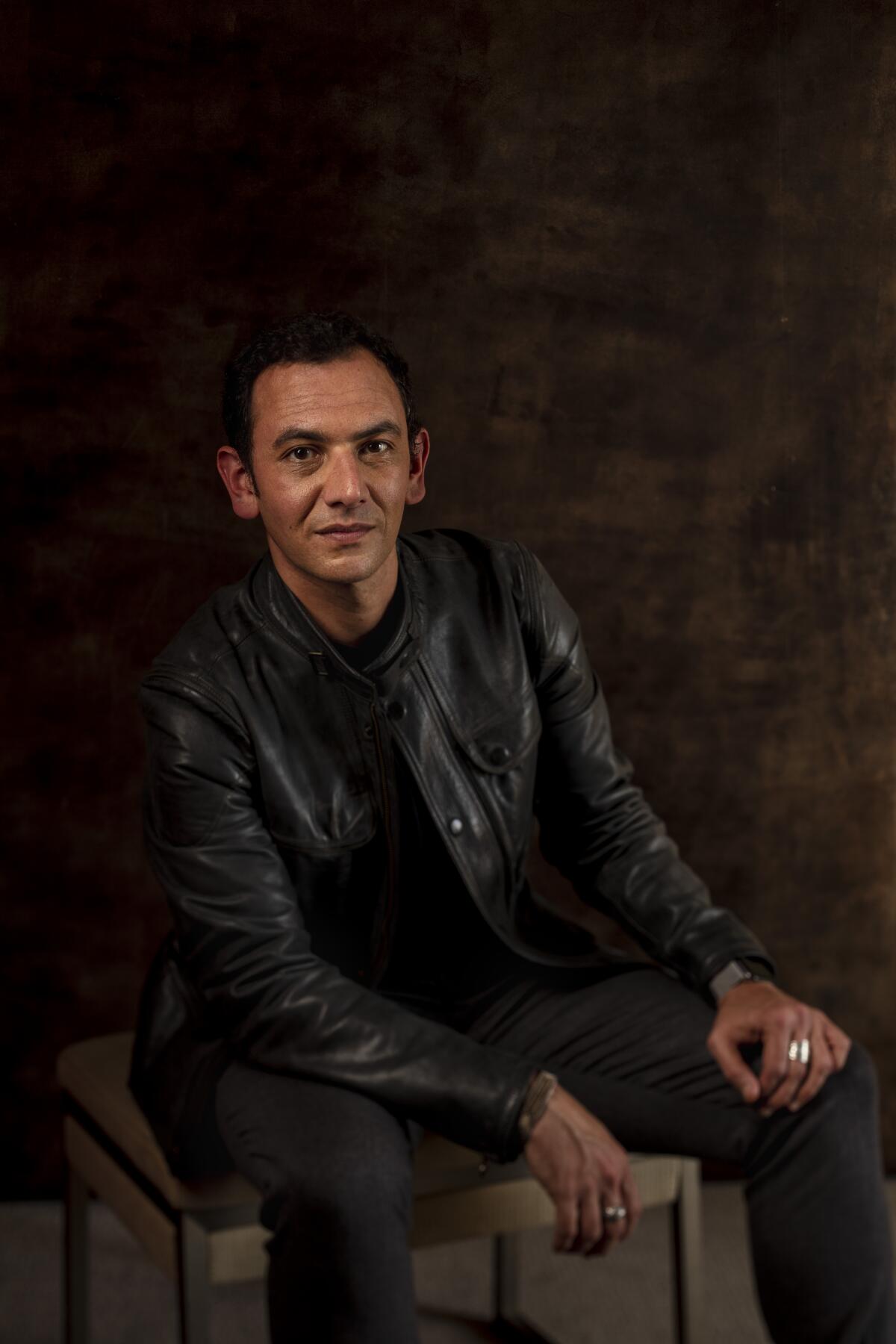 Screenwriter Roberto Bentivegna poses in a leather jacket sitting in a chair.