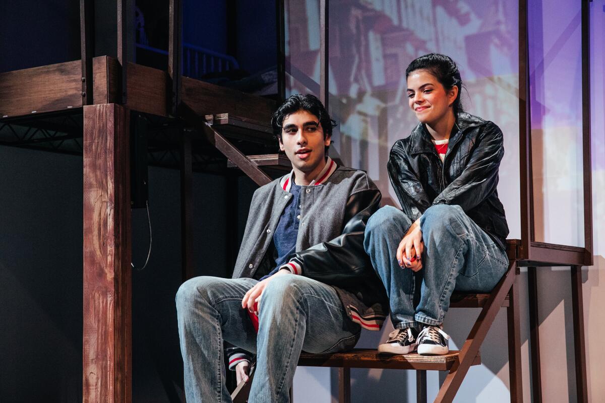 A young man and woman in jeans and sneakers sit onstage on a set of steps.