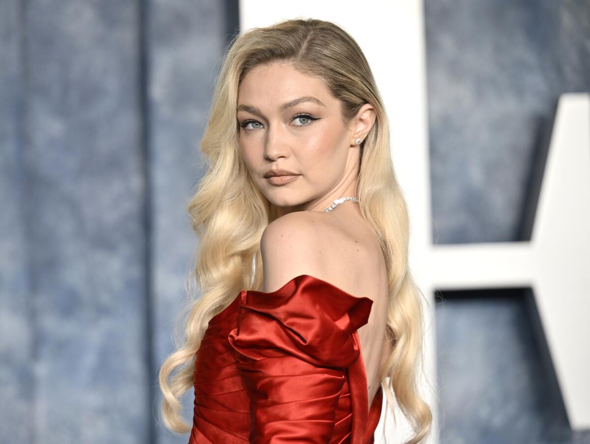 Gigi Hadid is looking over her shoulder while wearing a red strapless dress and jeweled necklace. 