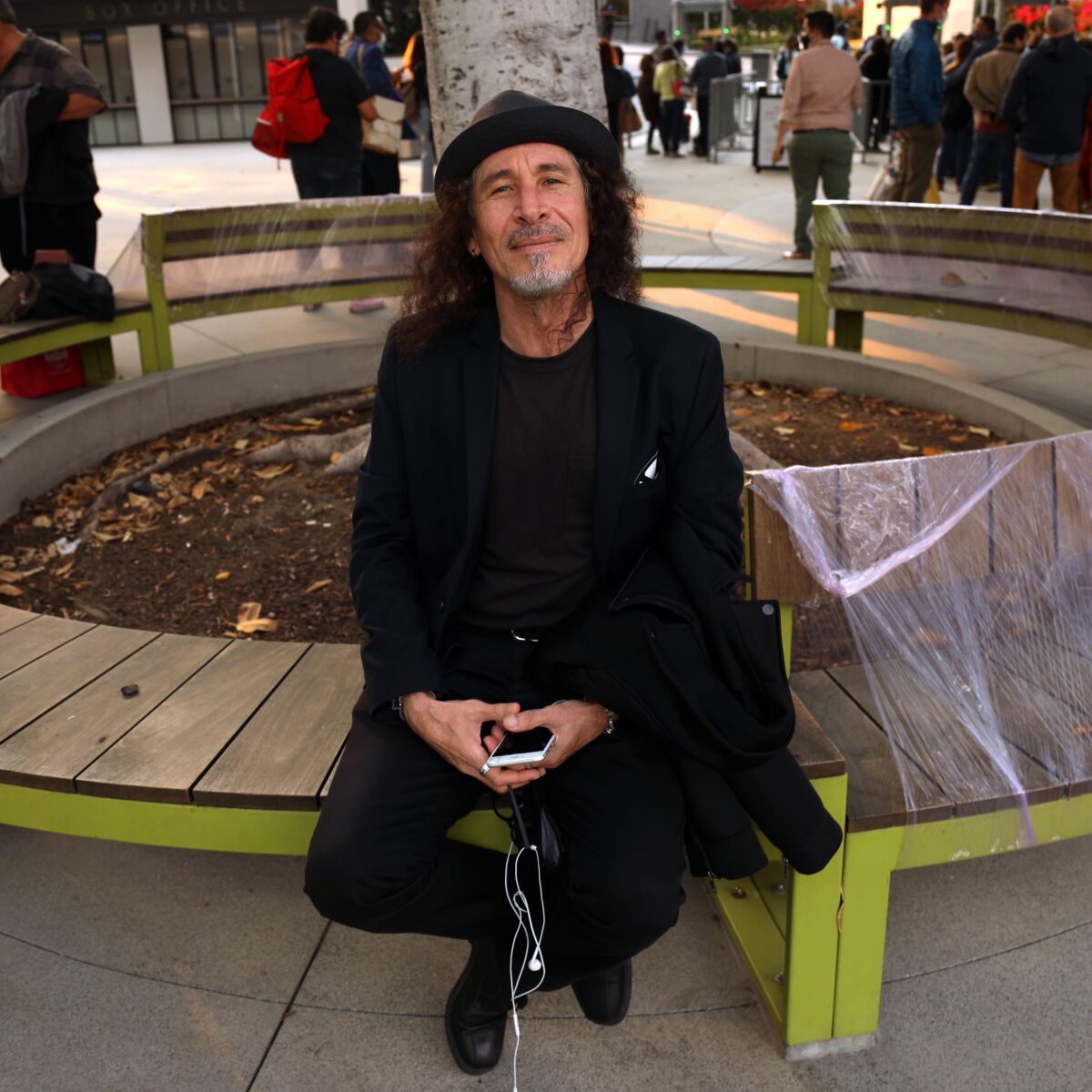 A man in a hat holds his phone as he poses for a photo seated on a bench.