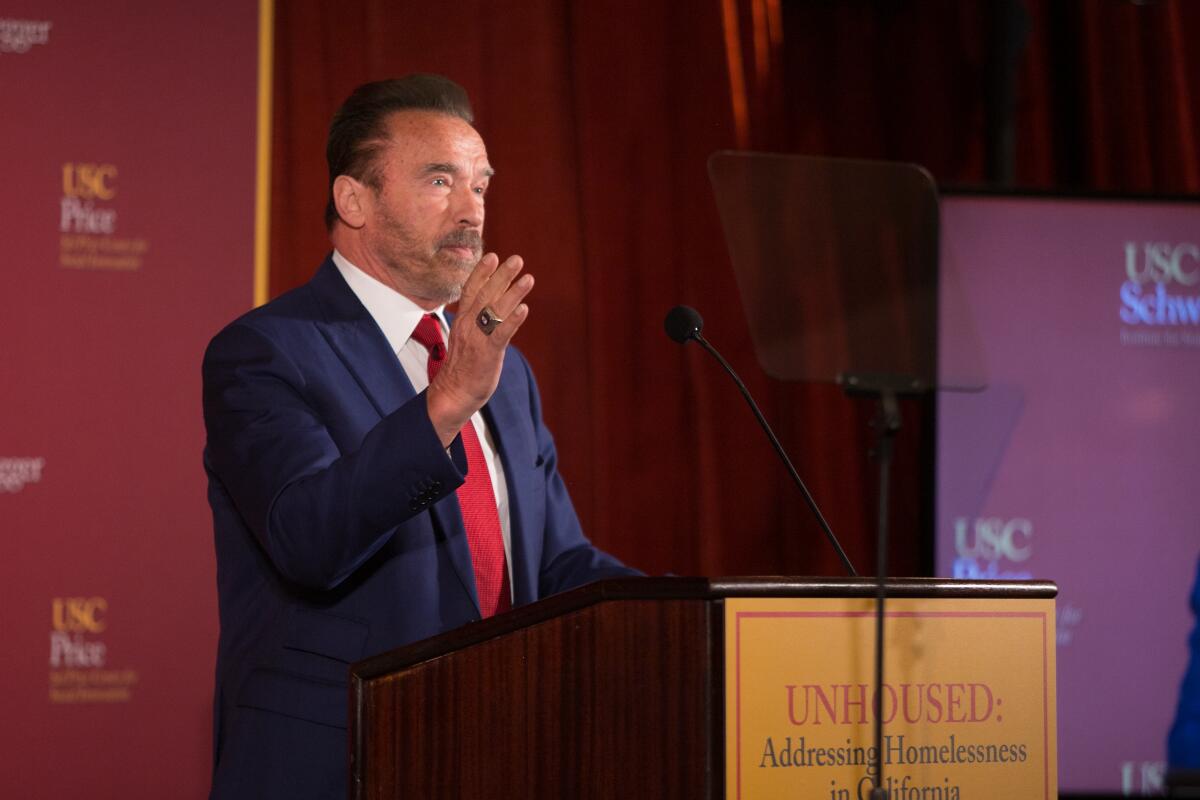 Former Gov. Arnold Schwarzenegger stands and gestures at a lectern with the USC logo in the background.