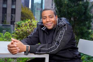 Kenan Thompson in a black jacket sitting on a metal chair with his hands clasped on a table