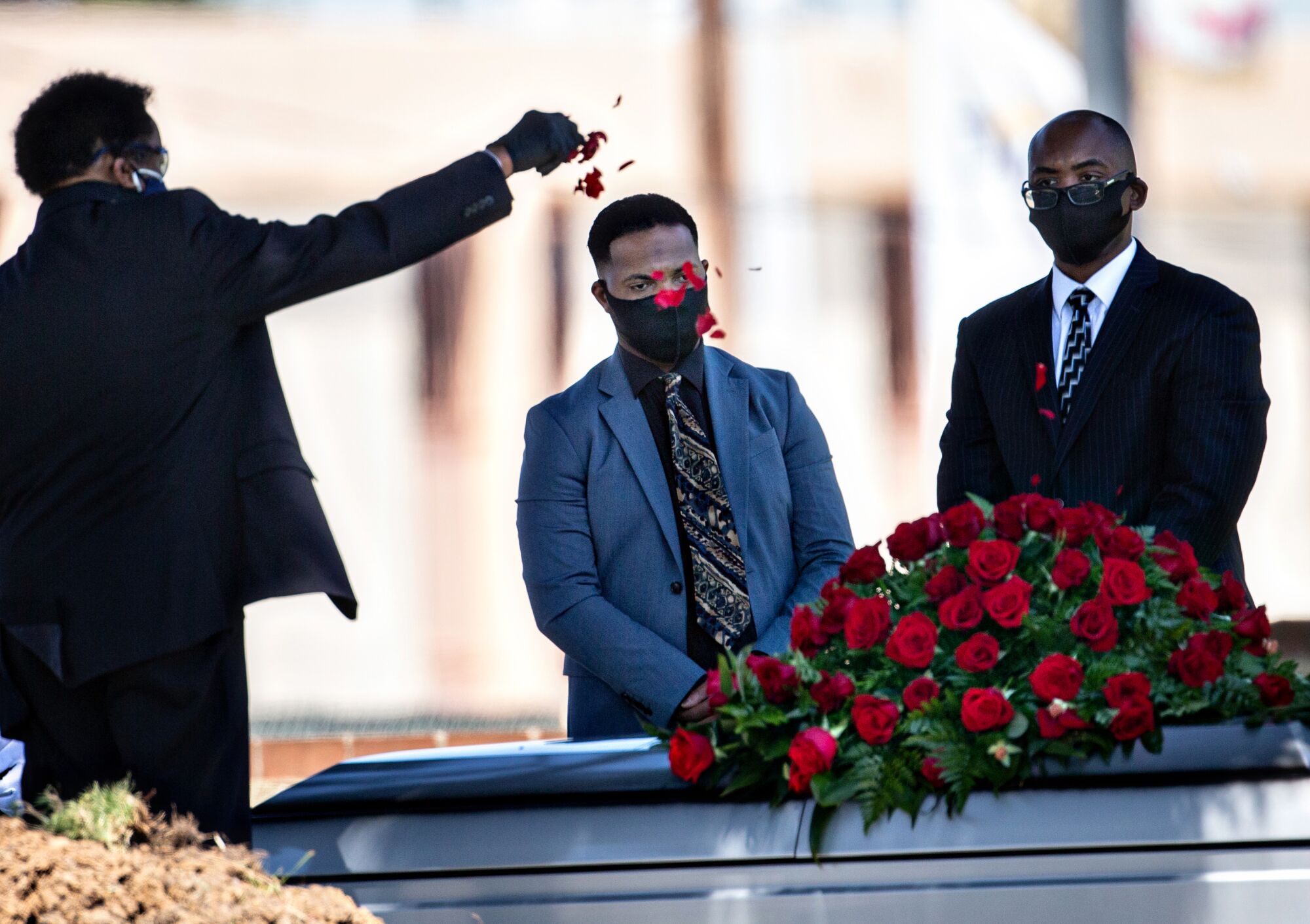A funeral director, next to mourners, sprinkles rose petals over a casket.
