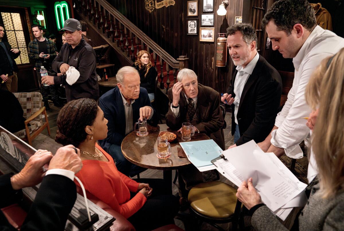 A group of people sit around a small table with glasses on it as two men stand holding scripts.