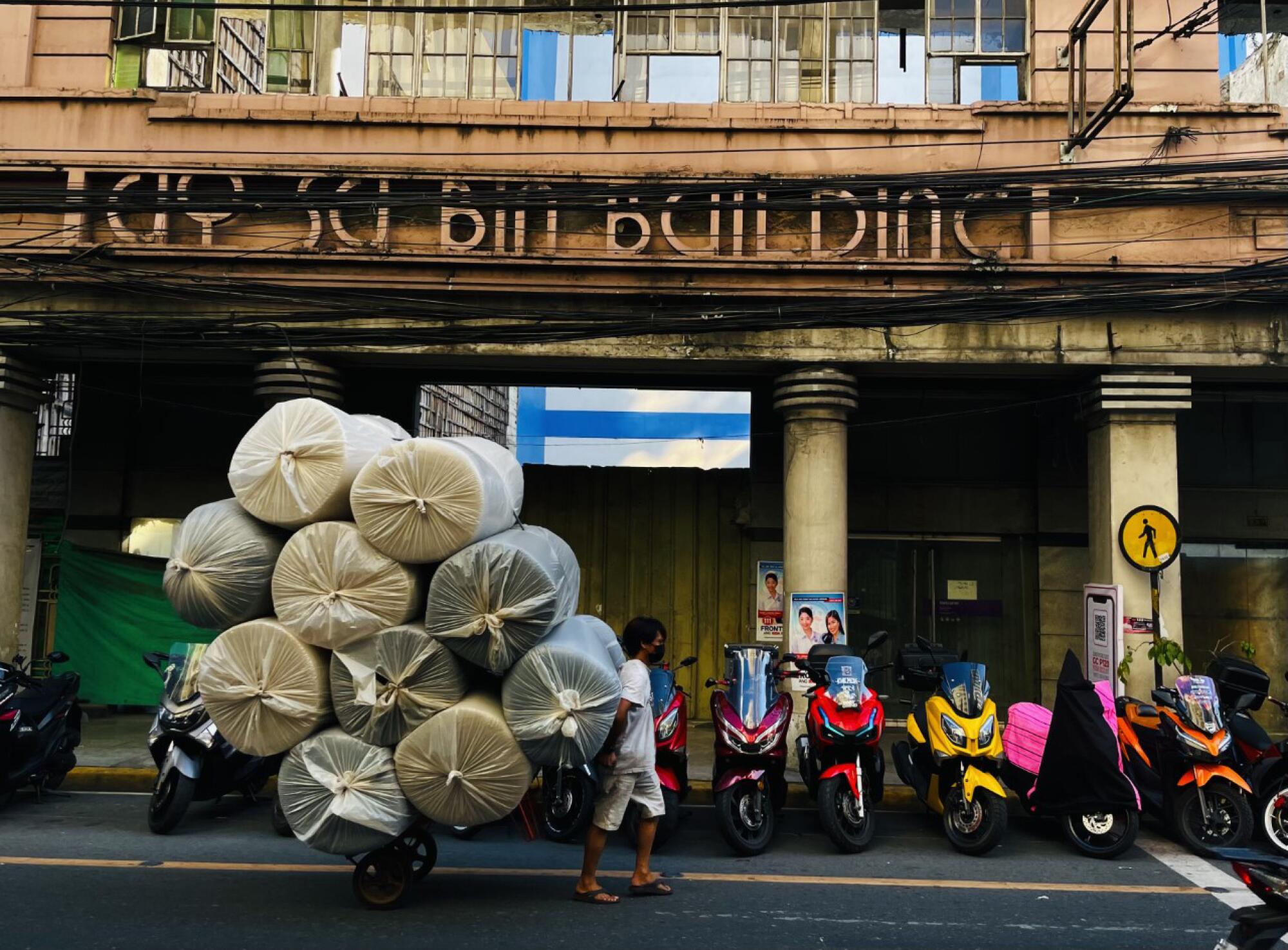 A laborer pulls a cart of rolled fabric past an old building with motor scooters parked in front.