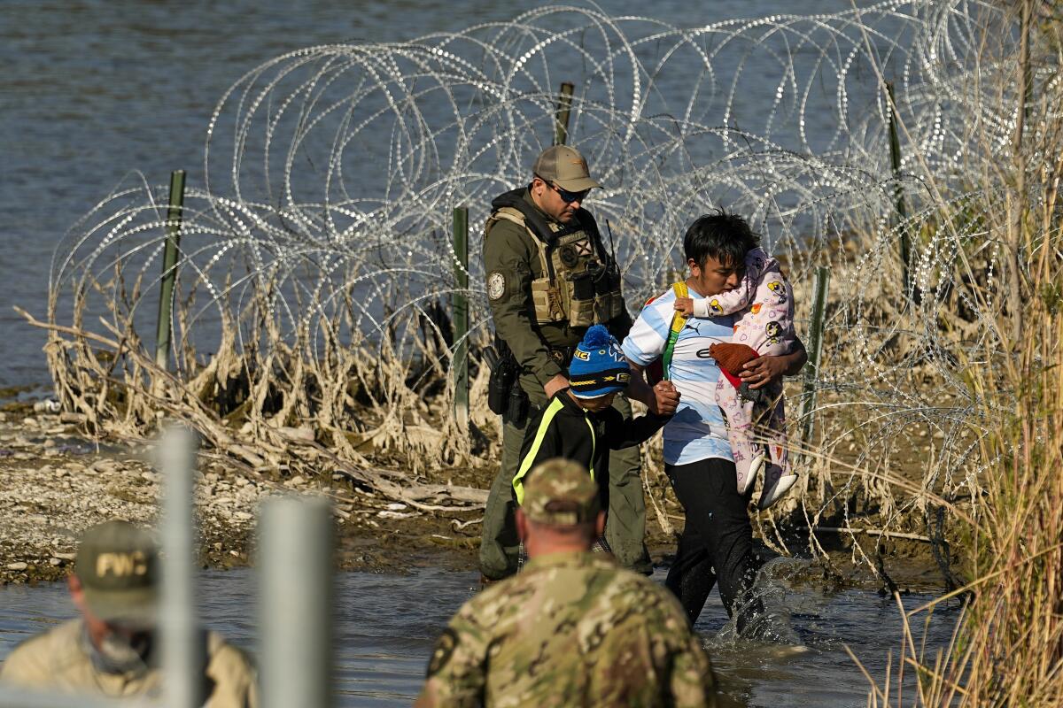 A person with two children is taken into custody at U.S.-Mexico border against a backdrop of razor wire.