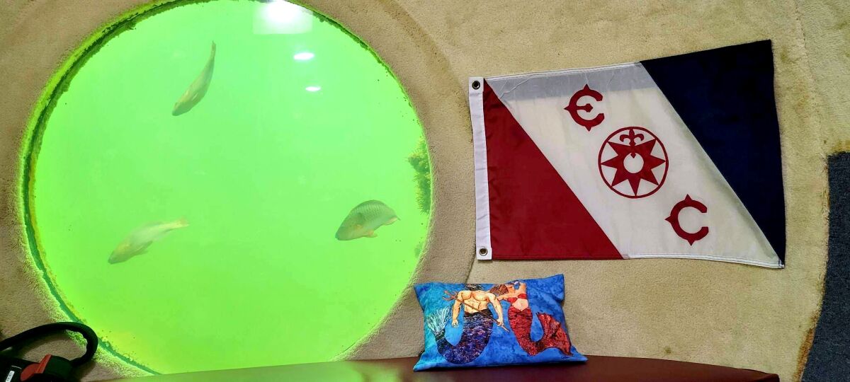 A room with a flag on the wall, a pillow, and a porthole framing bright-green water and three fish swimming by