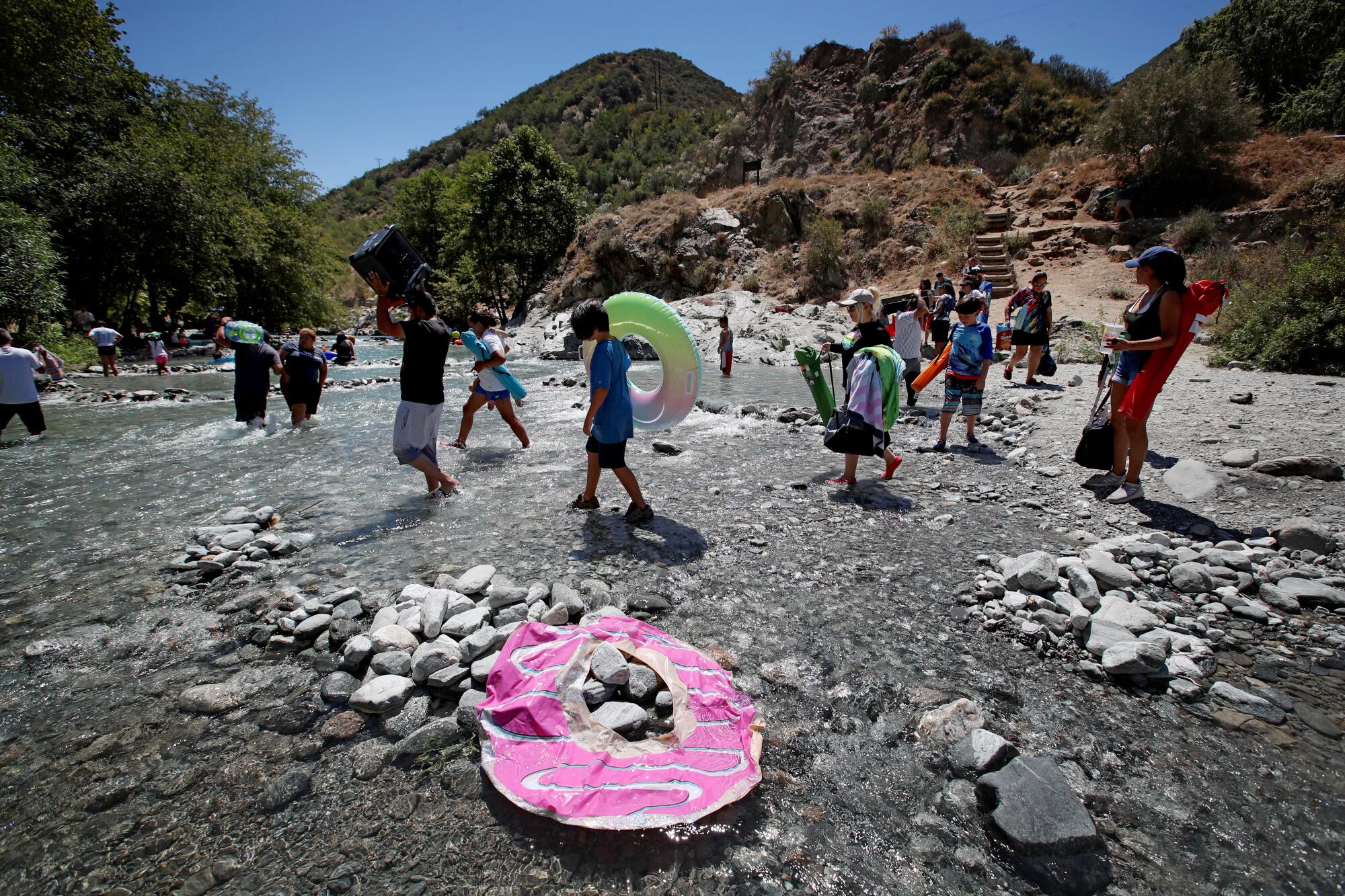 A deflated float toy lies on rocks as more than a dozen people wade into a shallow river.