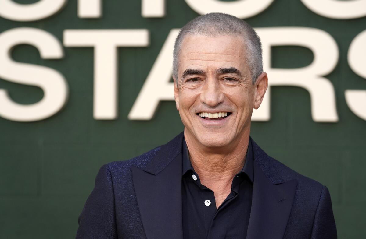Dermot Mulroney with buzzed hair in a blue blazer and dress shirt smiling against a green backdrop