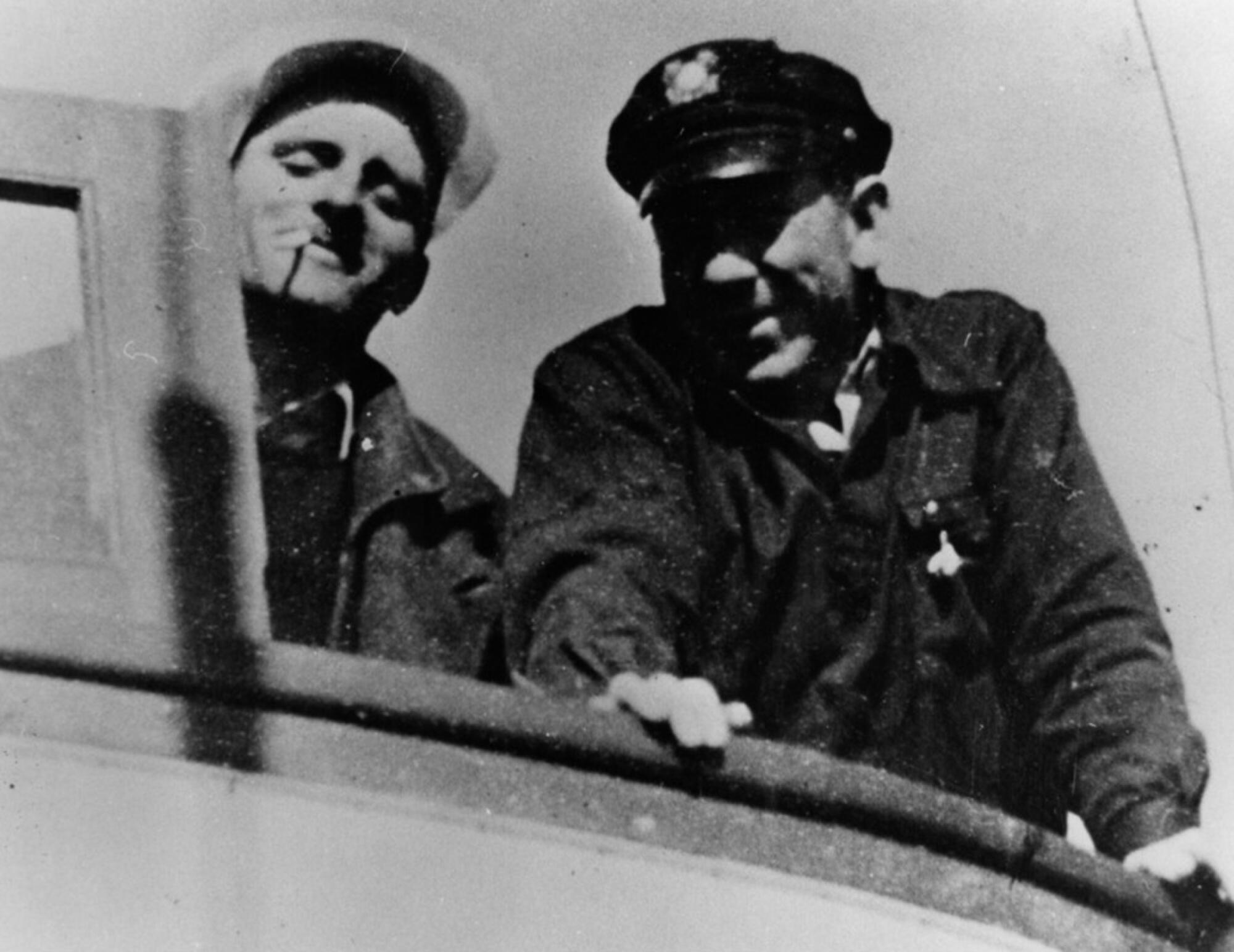 Two men, one with a cigarette in his mouth and the other in a sea captain's hat stand side by side on a boat