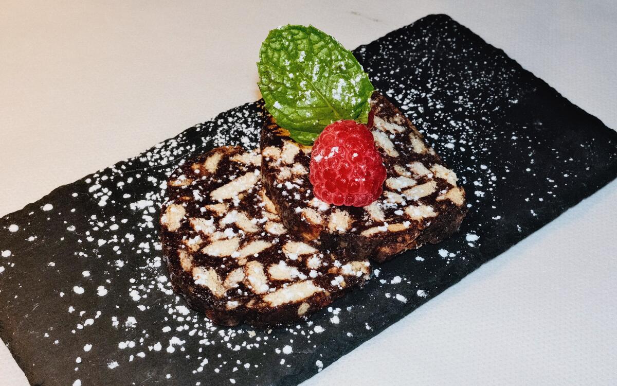 Two rounds of dark cocoa-powder cookies dotted with white cookie pieces on a black slate with powdered sugar and a raspberry