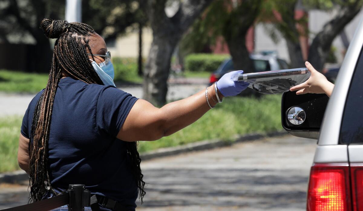 A woman in mask and gloves hands a computer to a motorist through a car window.