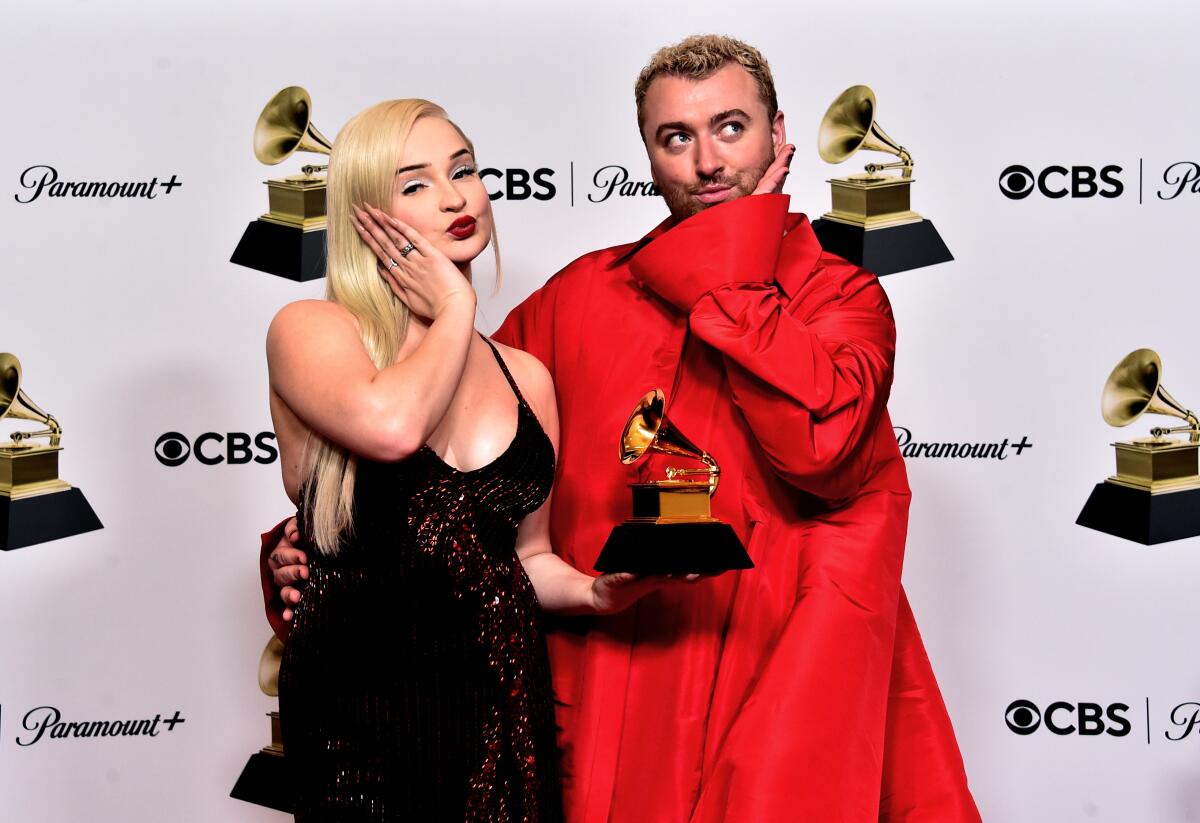 Kim Petras in a sparkling black dress, standing next to Sam Smith in a red outfit, and holding a Grammy Award