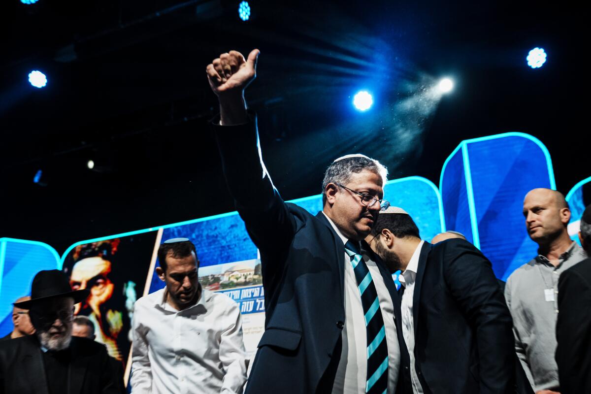 Itamar Ben-Gvir gives a thumbs-up and hugs a man as others stand by on a stage decorated with blue panels