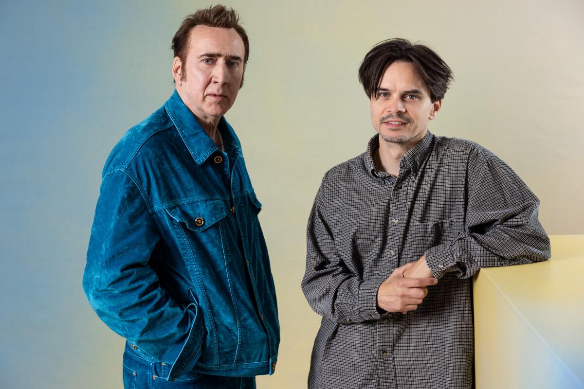 Nicolas Cage wears a denim jacket while standing with Kristoffer Borgli, who rests his arm on a tall block for a portrait.
