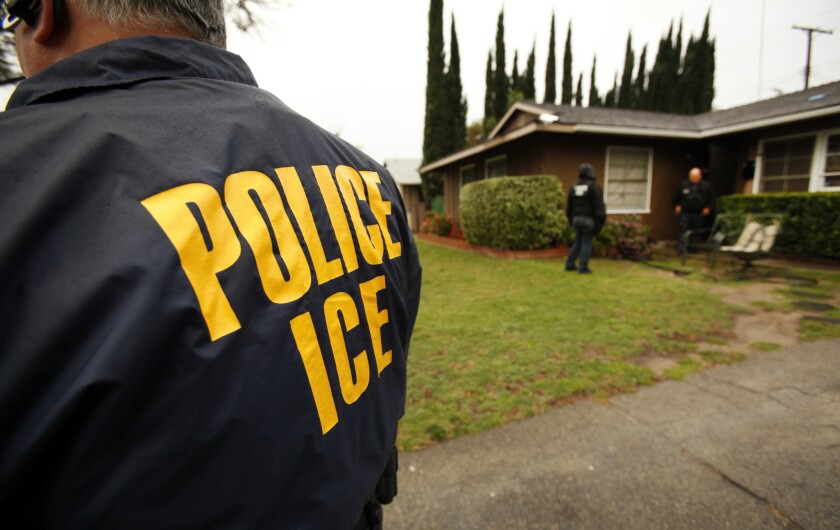 A man stands with his back to the camera in front of a one-story house. He is wearing a jacket that reads "POLICE" and "ICE."