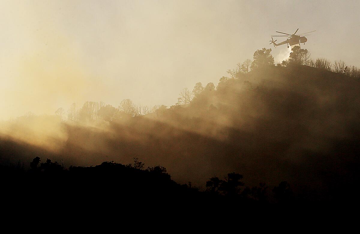 A helicopter flies above a smoky ridge dropping water.