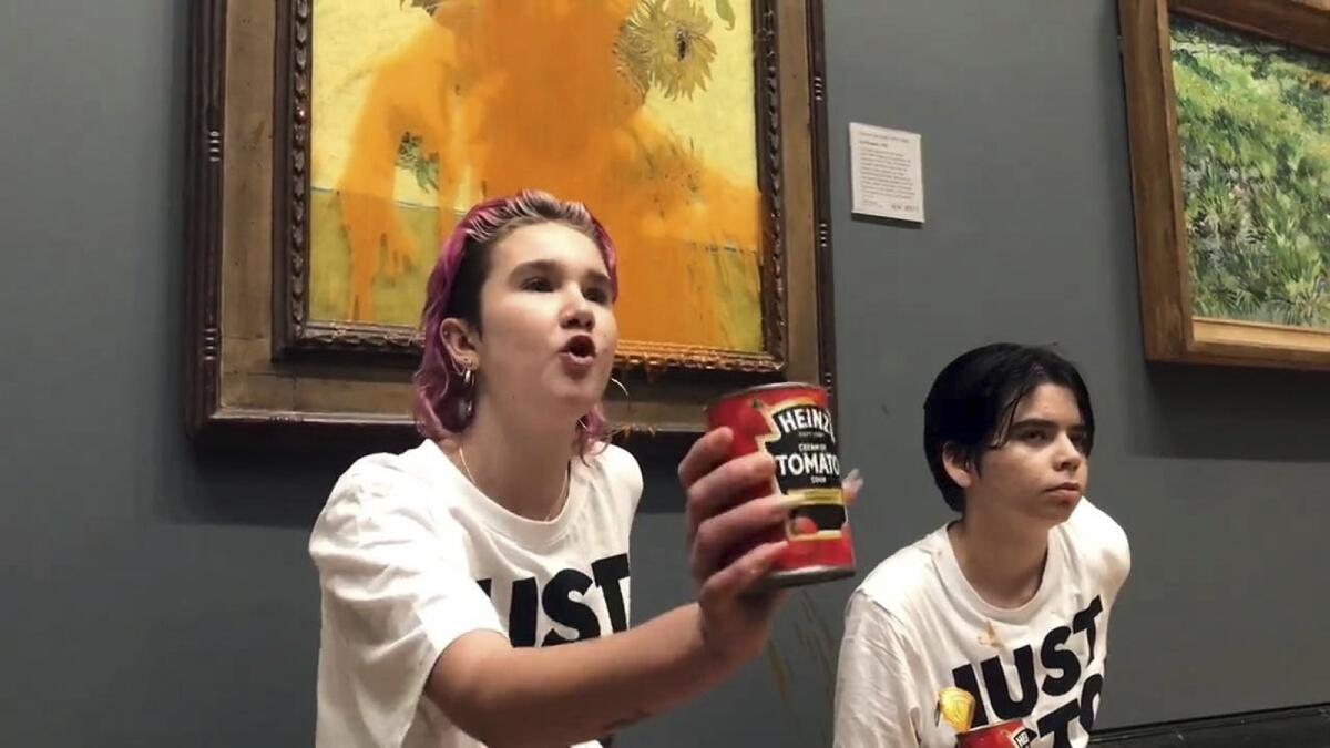 Two young people hold a can of tomato soup up in front of Vincent Van Gogh's painting 'Sunflowers.'