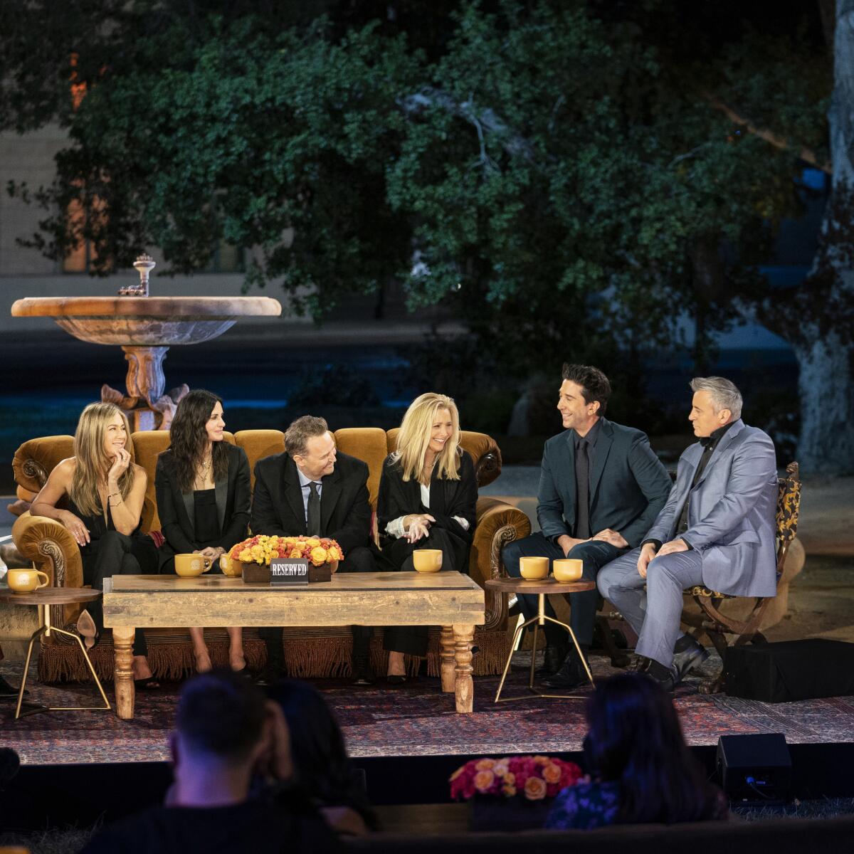 Six familiar actors in semi-formal attire sit on a sofa and cushy chair behind a coffee table and in front of a fountain