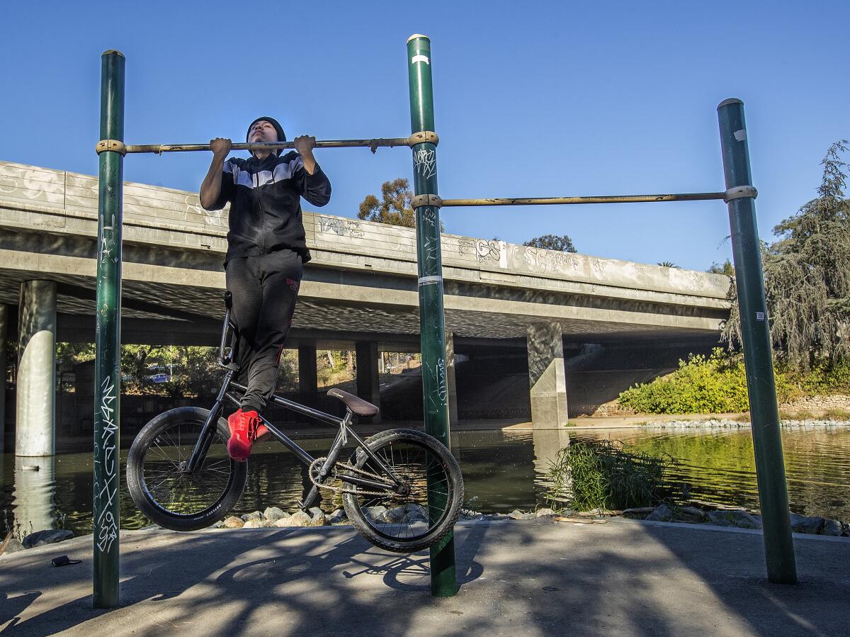 With a freeway overpass looming in the back, a teen lifts himself on a pull-up bar with his bicycle entwined in his feet.