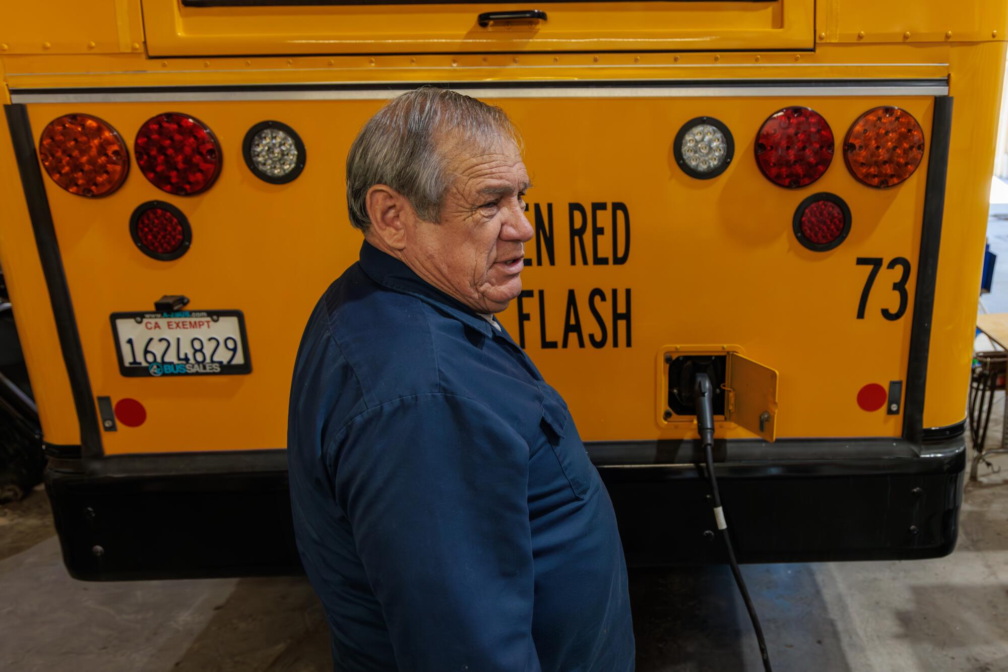 A man stands at the rear of school bus where a charging cable is connected.
