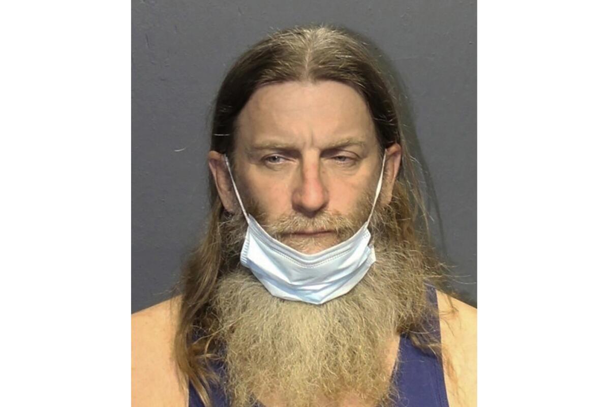 A mug shot of a man with long brown hair and graying mustache and beard and blue mask on his chin 