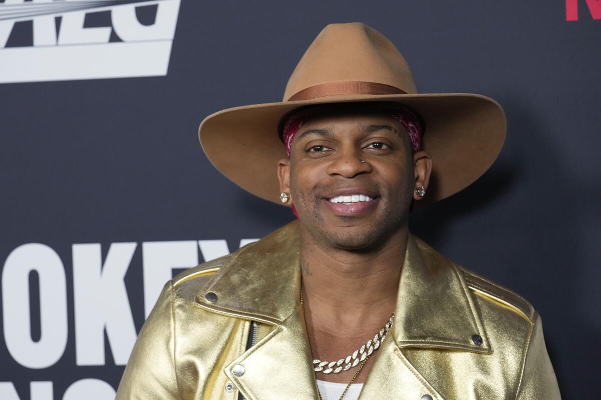 Jimmie Allen wears a gold leather jacket with a red bandana under a beige cowboy hat