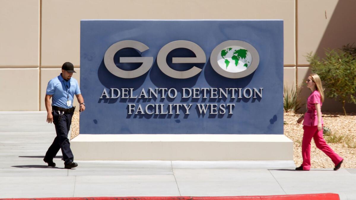 Two people walk past a sign for a GEO Group immigrant detention center