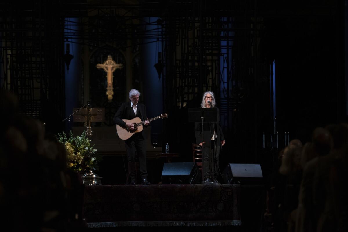 A man holds a guitar next to a woman standing at a microphone onstage in a darkened space.