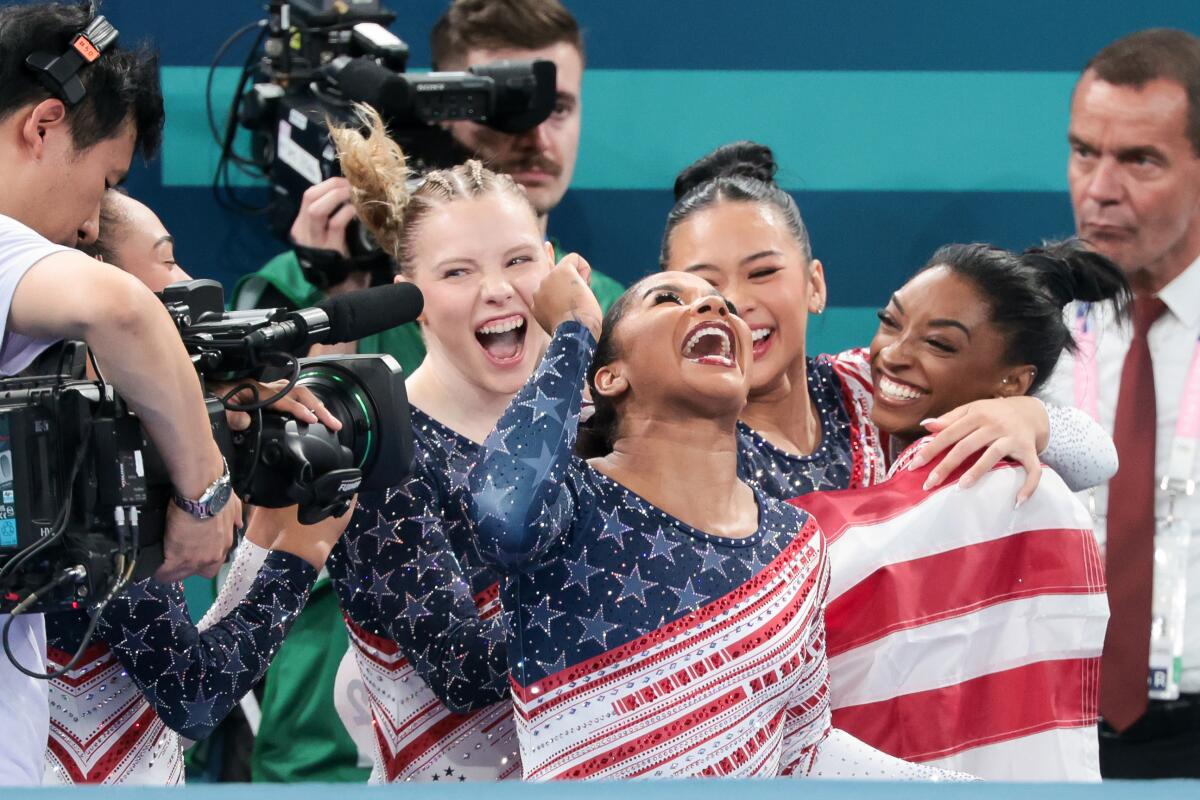 After winning a gold, Jordan Chiles stands next to her teammates and laughs while wrapped in an American flag.
