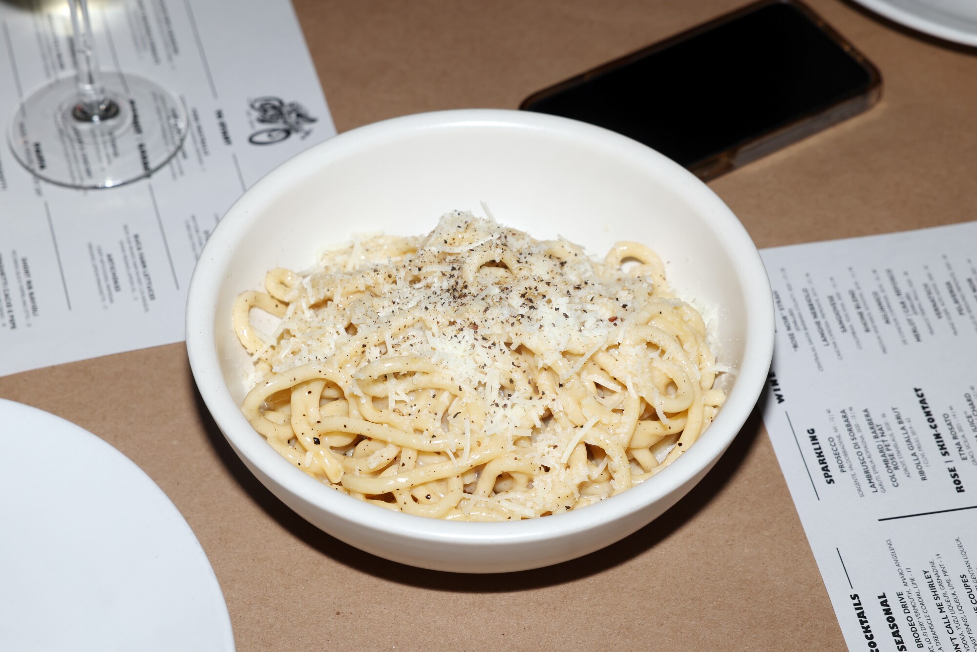 A bowl of pasta sits on a table amid menus, wine glasses and plates.