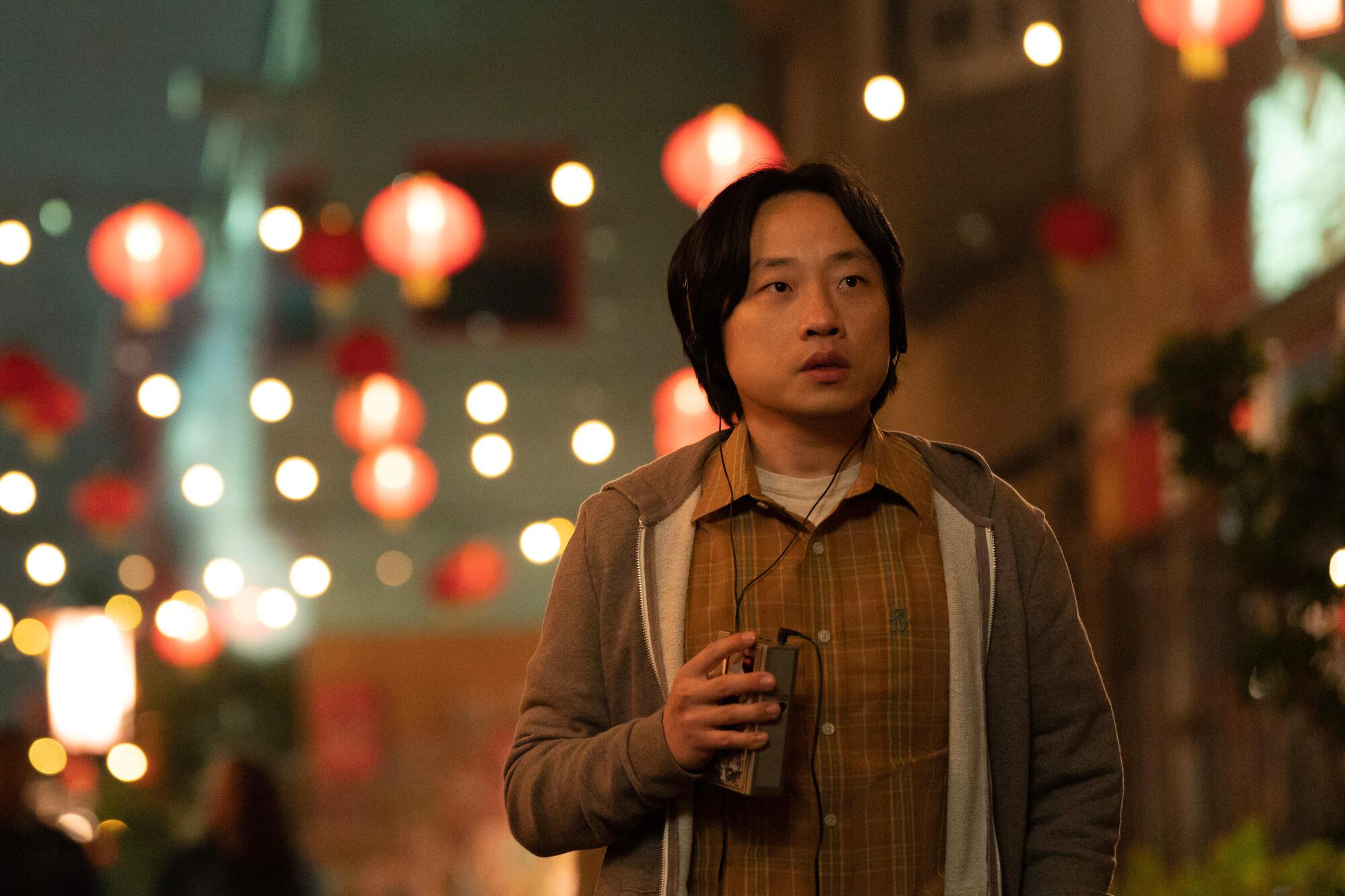 A man wearing headphones and holding an audio device stands outside under strings of paper lanterns.