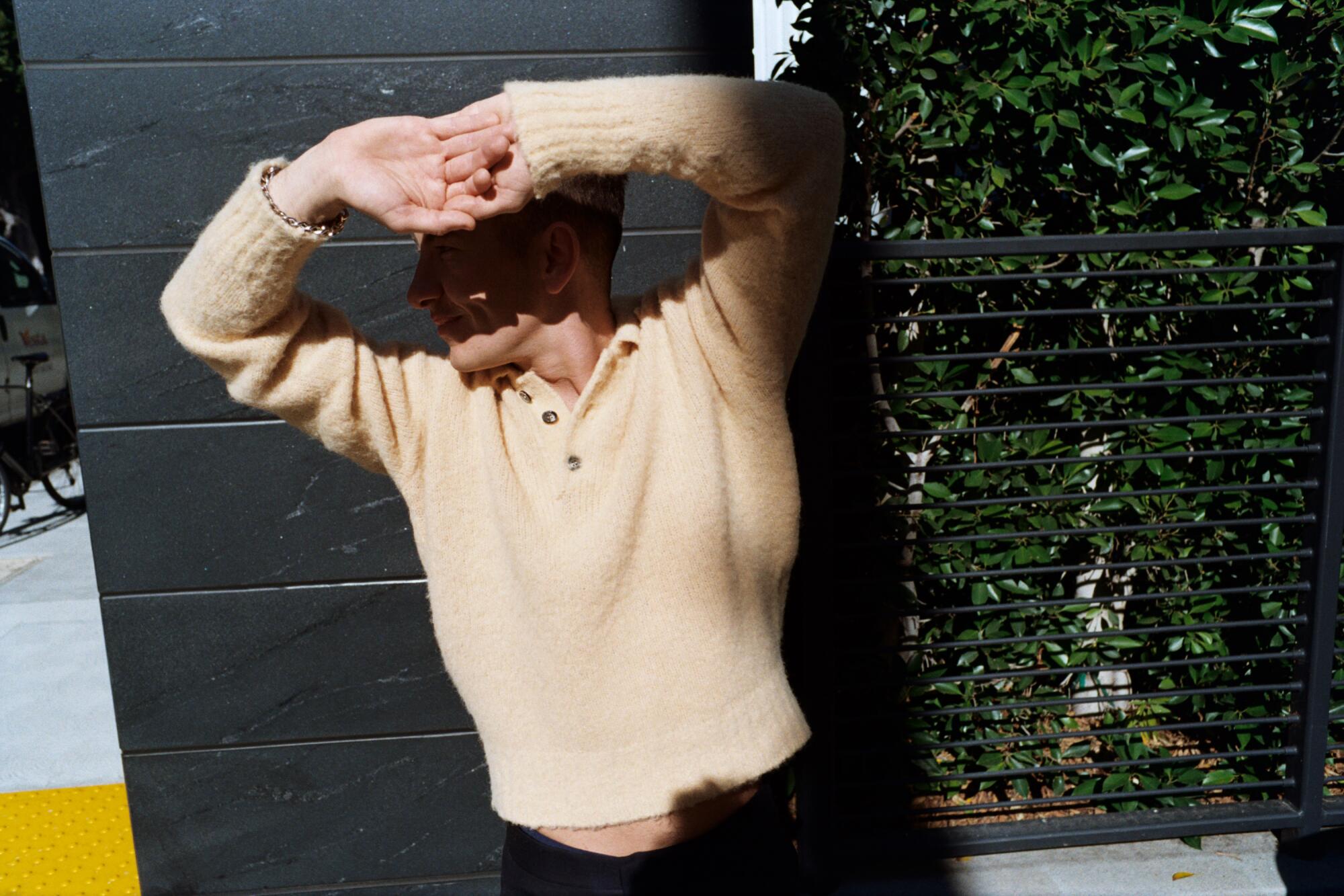 A man in a tan sweater shields his eyes from the sun with his hands, casting a dark shadow on his face.