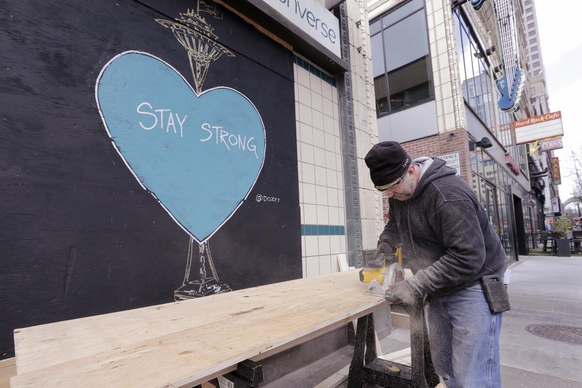 A carpenter cuts plywood to cover a closed store where artwork in support of people affected by the coronavirus already covers an adjacent panel in downtown Seattle on  Thursday. 