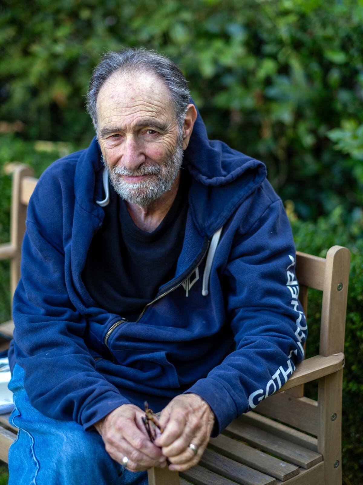 An older man in a hoodie leans forward with his hands together.